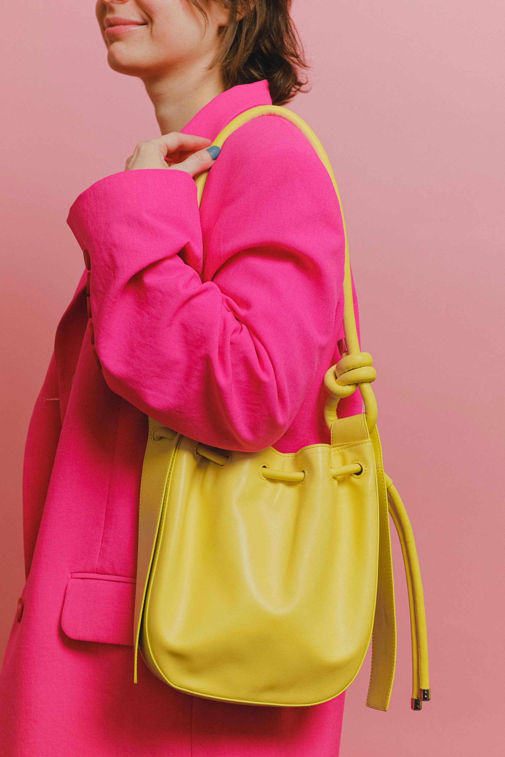 Fashionable woman in pink blazer holding yellow shoulder bag against pink background.