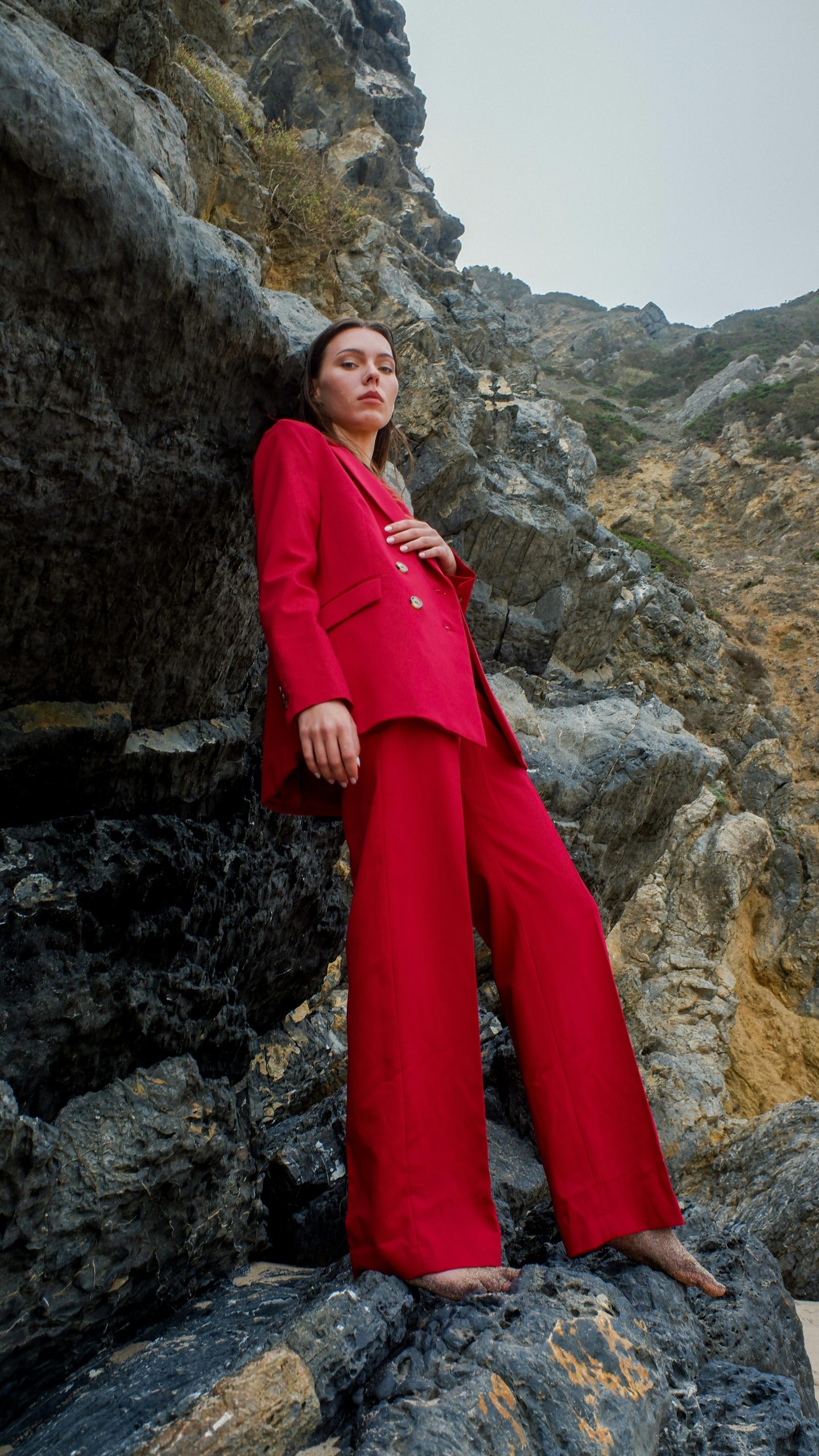 A fashionable woman in a striking red suit leans against rocky terrain, showcasing modern elegance.